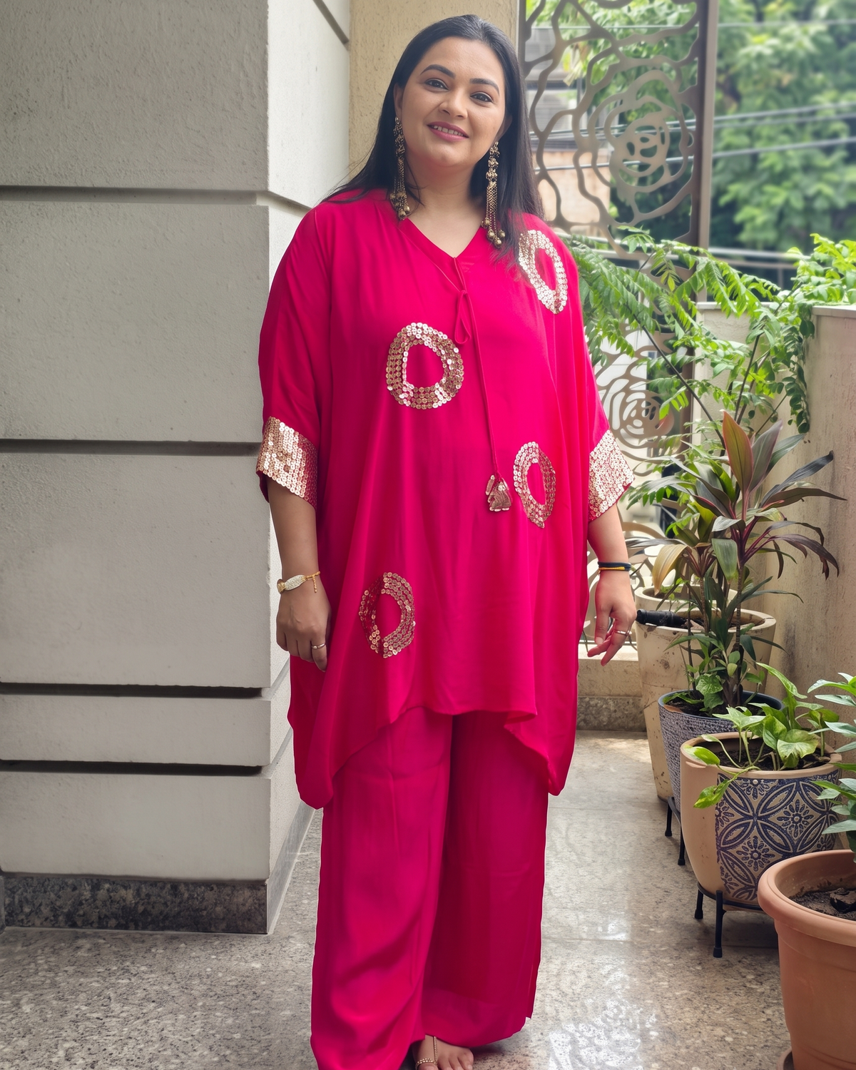 A woman modeling a bright pink two-piece kurta and trouser set with gold circular and border patterns, standing on an outdoor balcony.